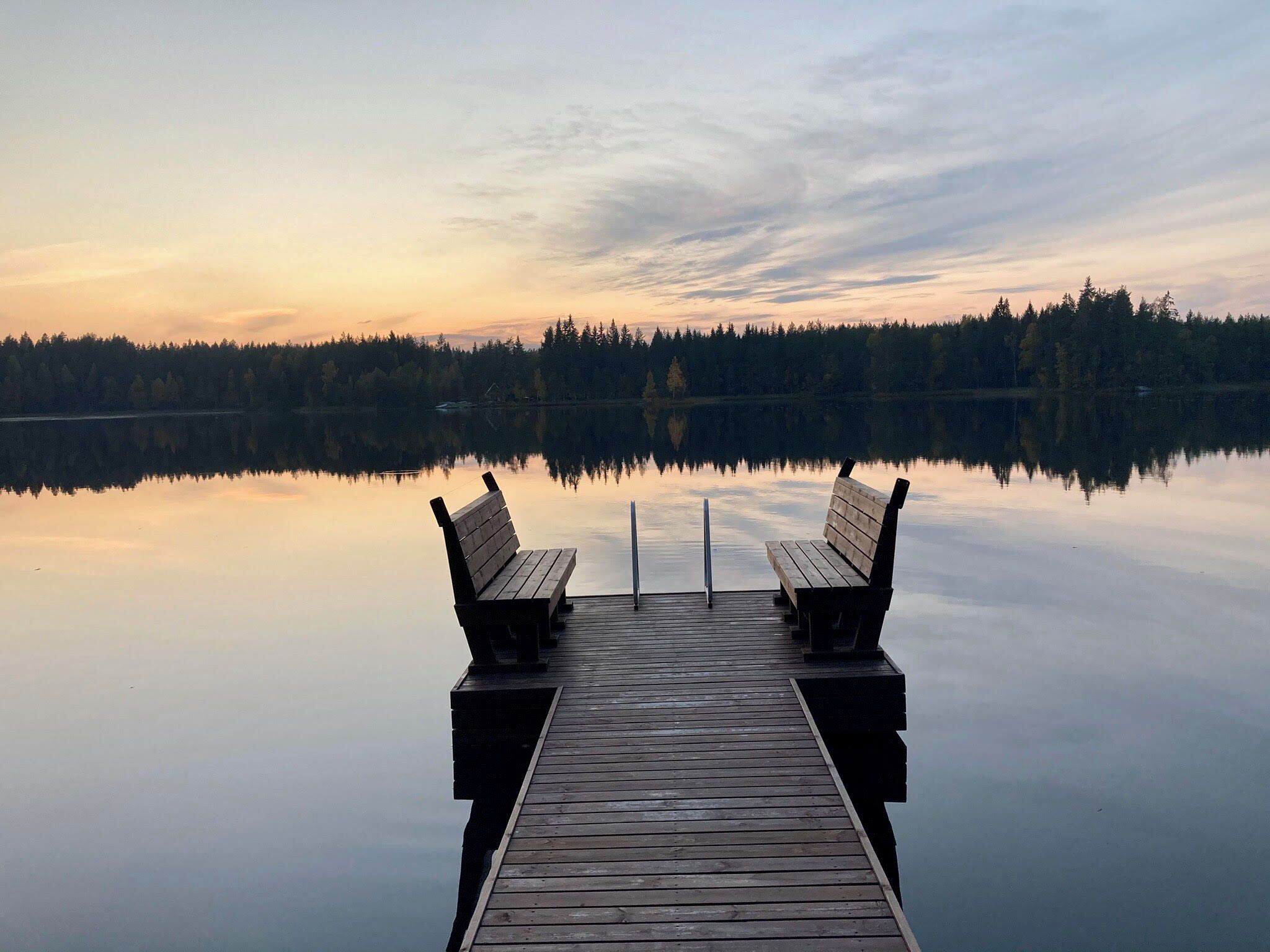 A wooden dock extending into calm water with two benches facing each other, surrounded by a forested shoreline.