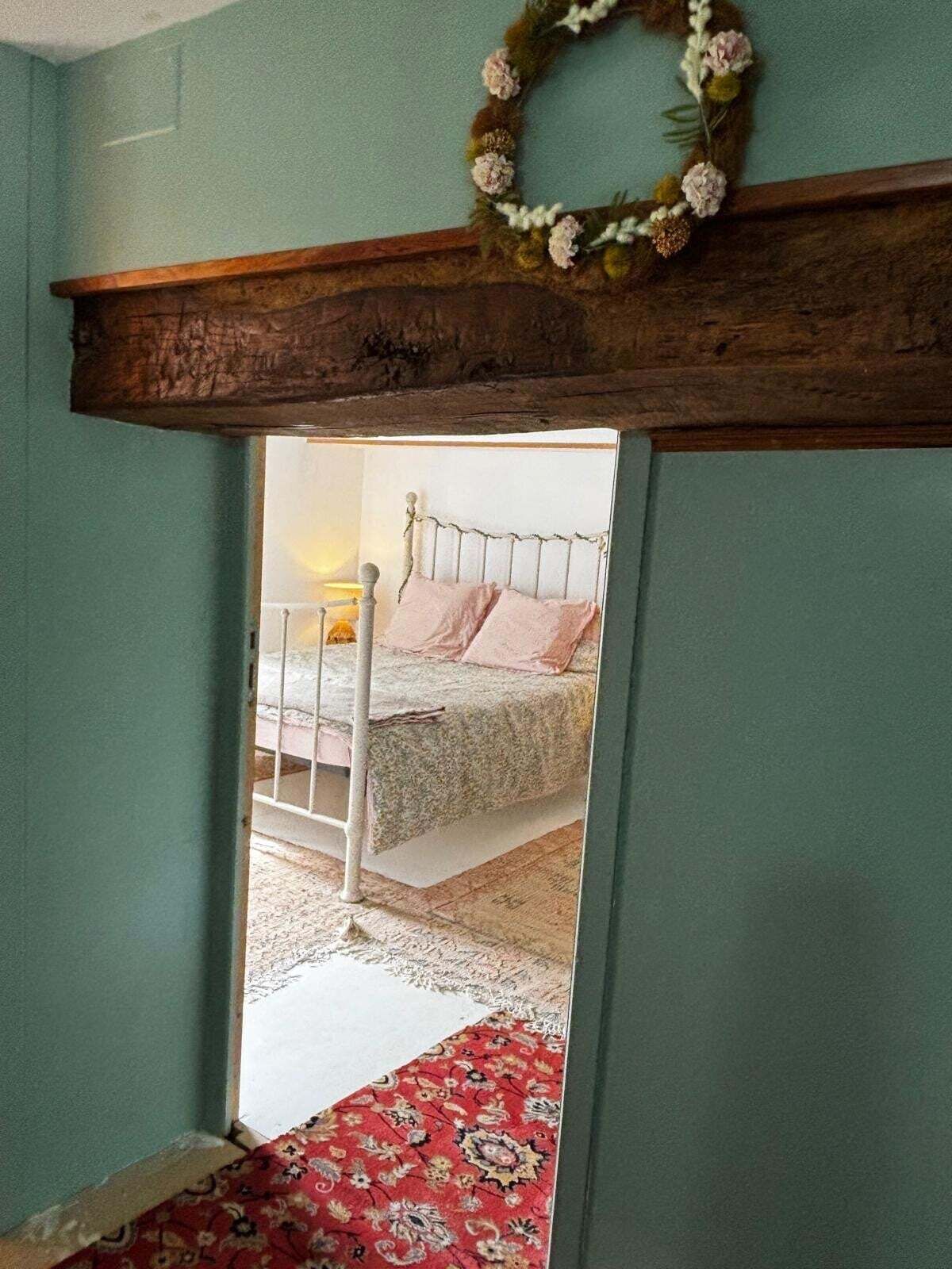 A bedroom with a bed, a wooden headboard, a red patterned rug, and a floral wreath on the wall.