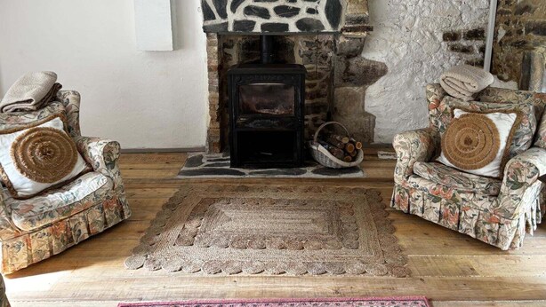 Una sala de estar acogedora con una chimenea de piedra, dos sillones con estampado floral, un tapiz tejido y una cesta de leña.