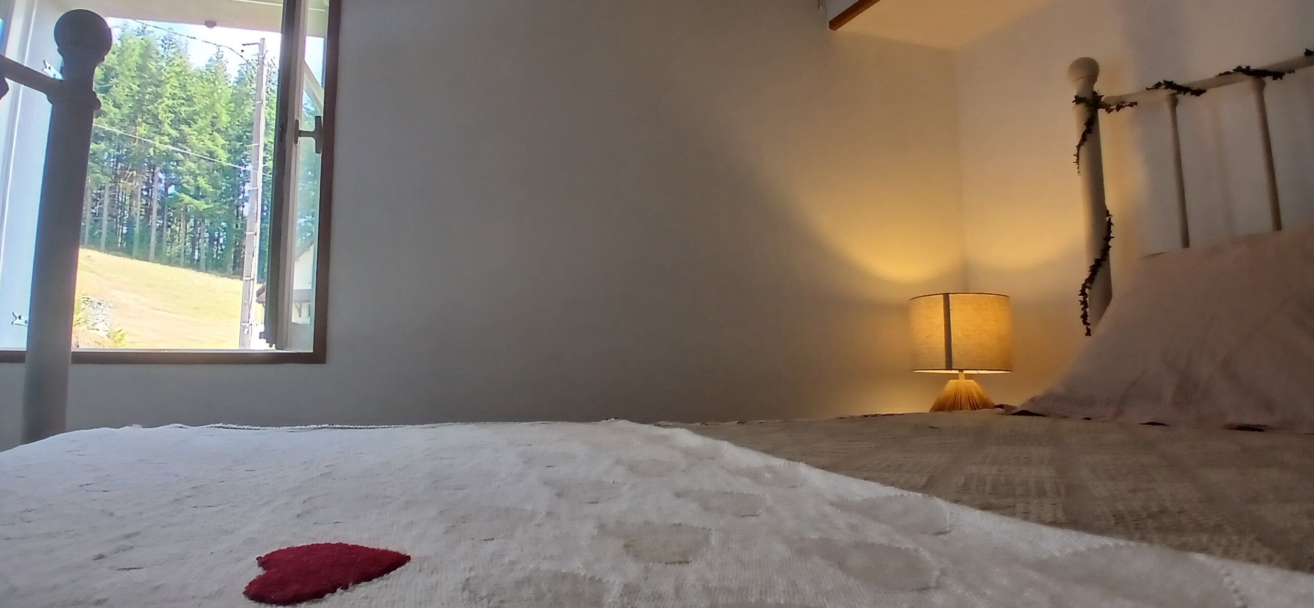 Ein Schlafzimmer mit Bett, Fenster, Lampe und Blick auf Bäume.