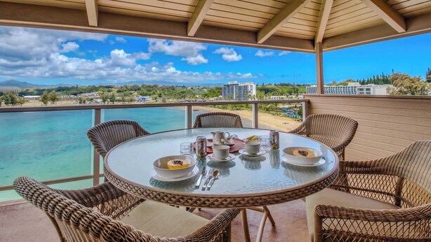 Une table ronde en verre, des chaises en osier, une vue sur la plage et un ciel dégagé.