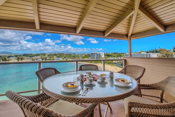 A round glass table with wicker chairs, a view of a beach, and a clear sky.