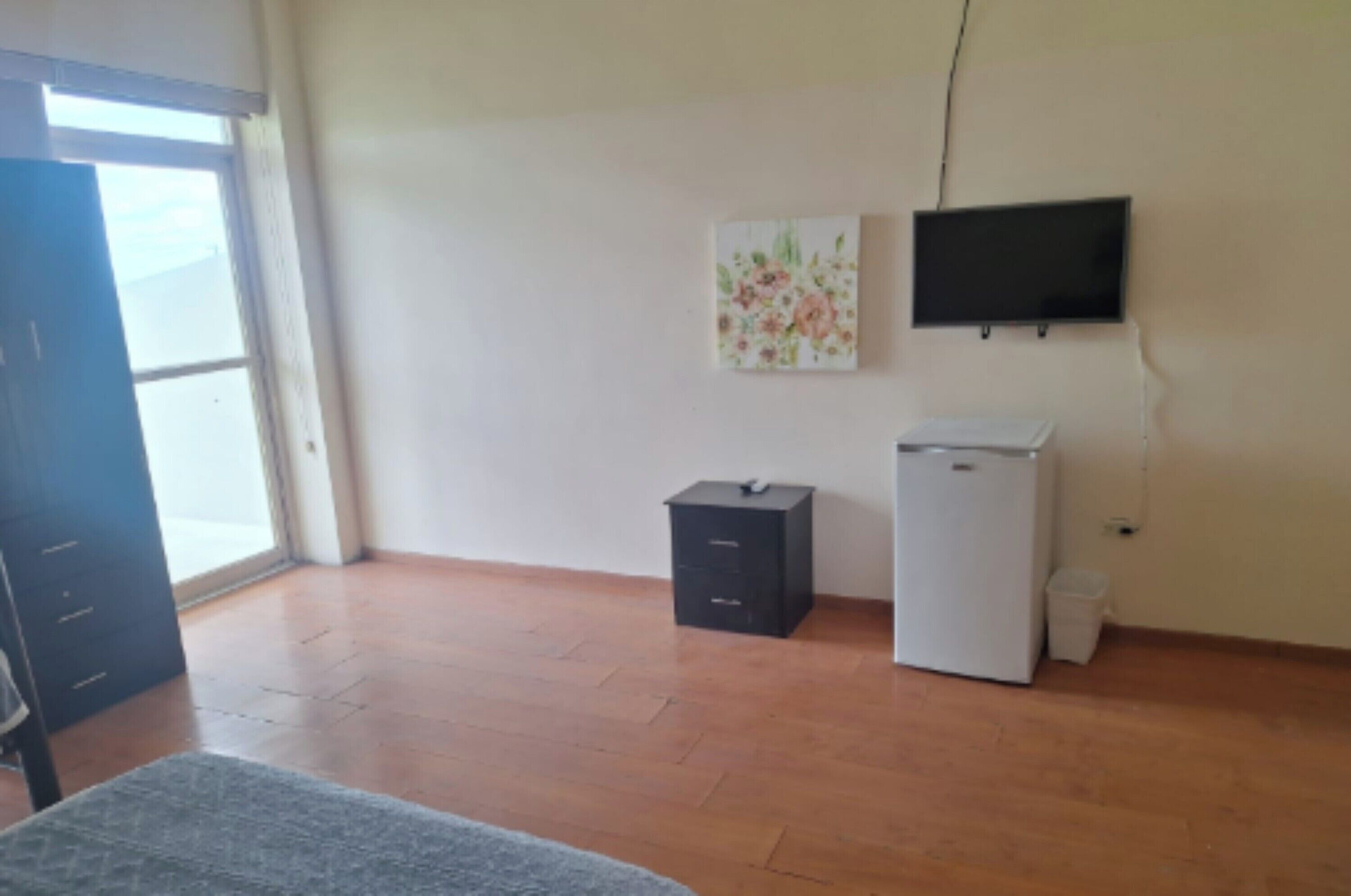 Een kamer met een groot raam, een wandgemonteerde flatscreen televisie, een kleine koelkast en een kast.
