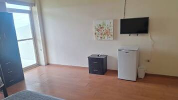 Een kamer met een groot raam, een wandgemonteerde flatscreen televisie, een kleine koelkast en een kast.
