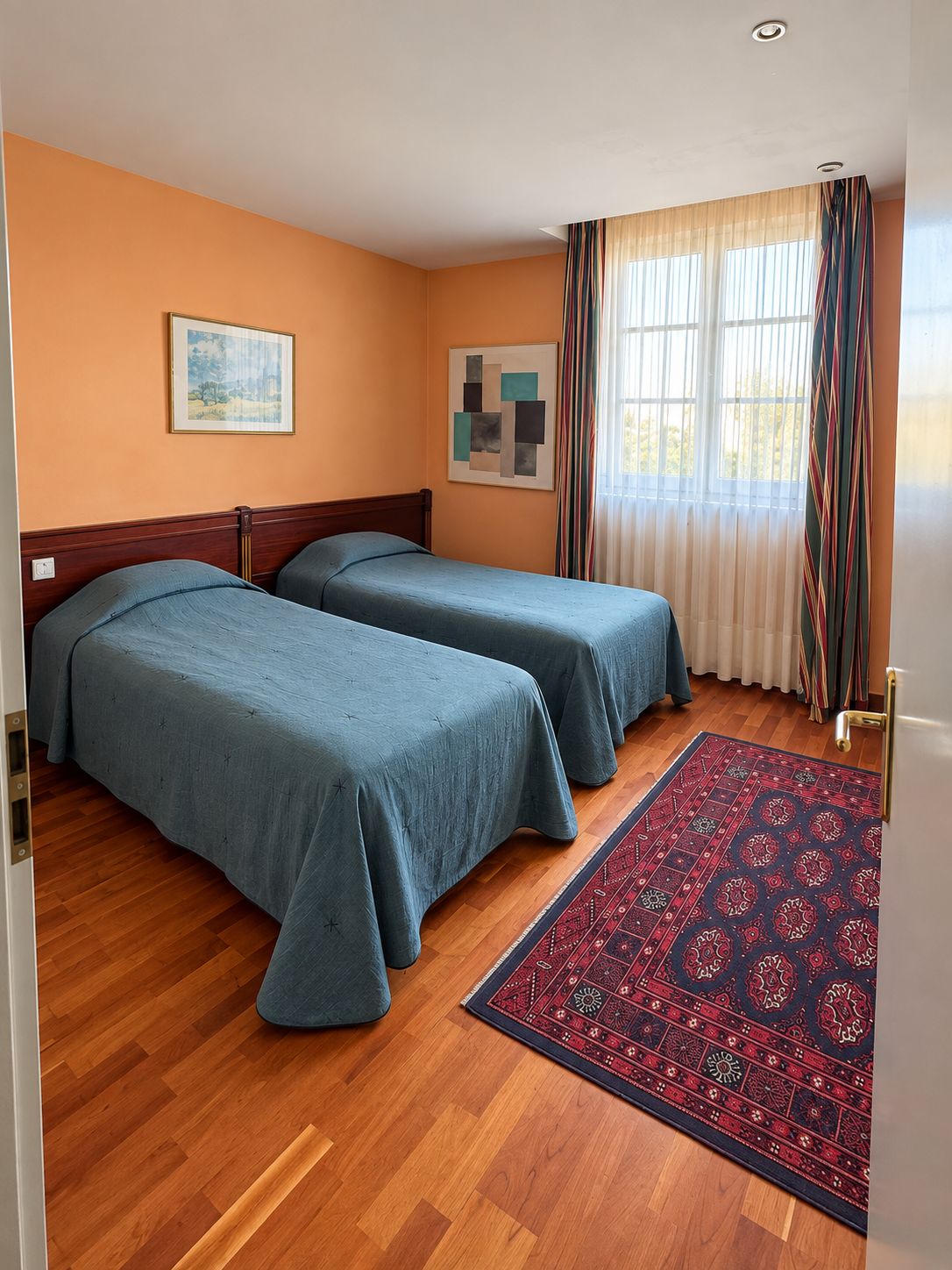 A room with two beds, a wooden floor, a patterned rug, and a window with curtains.