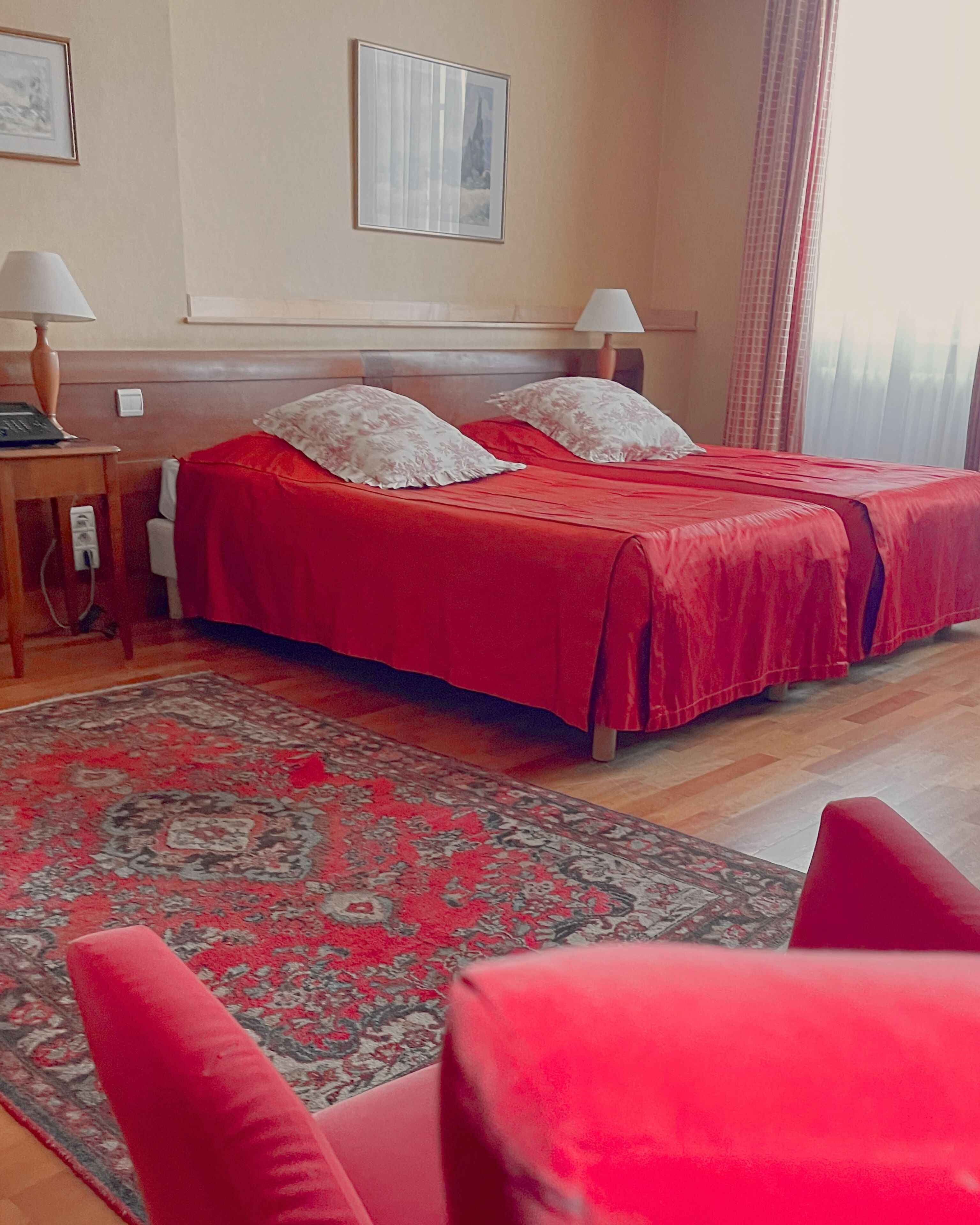 Une chambre d’hôtel avec deux lits, une moquette rouge, un plancher en bois, une table de chevet, une lampe et une fenêtre avec des rideaux.