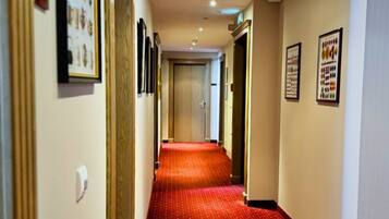 Pasillo de hotel con alfombra roja, cuadros en las paredes y una puerta al fondo.