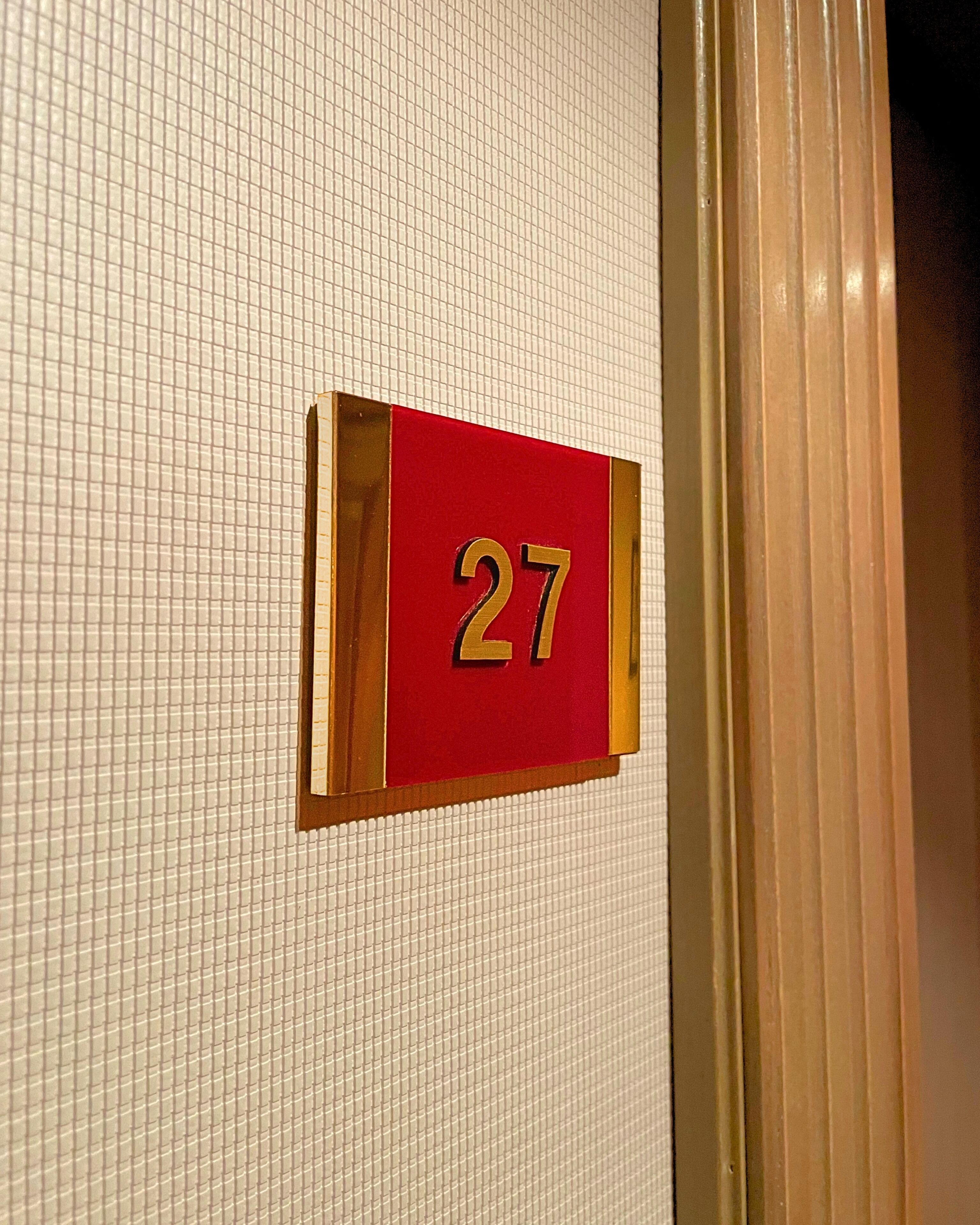 Een close-up van een hotelkamer nummerbordje, '27', bevestigd aan een betegelde muur.