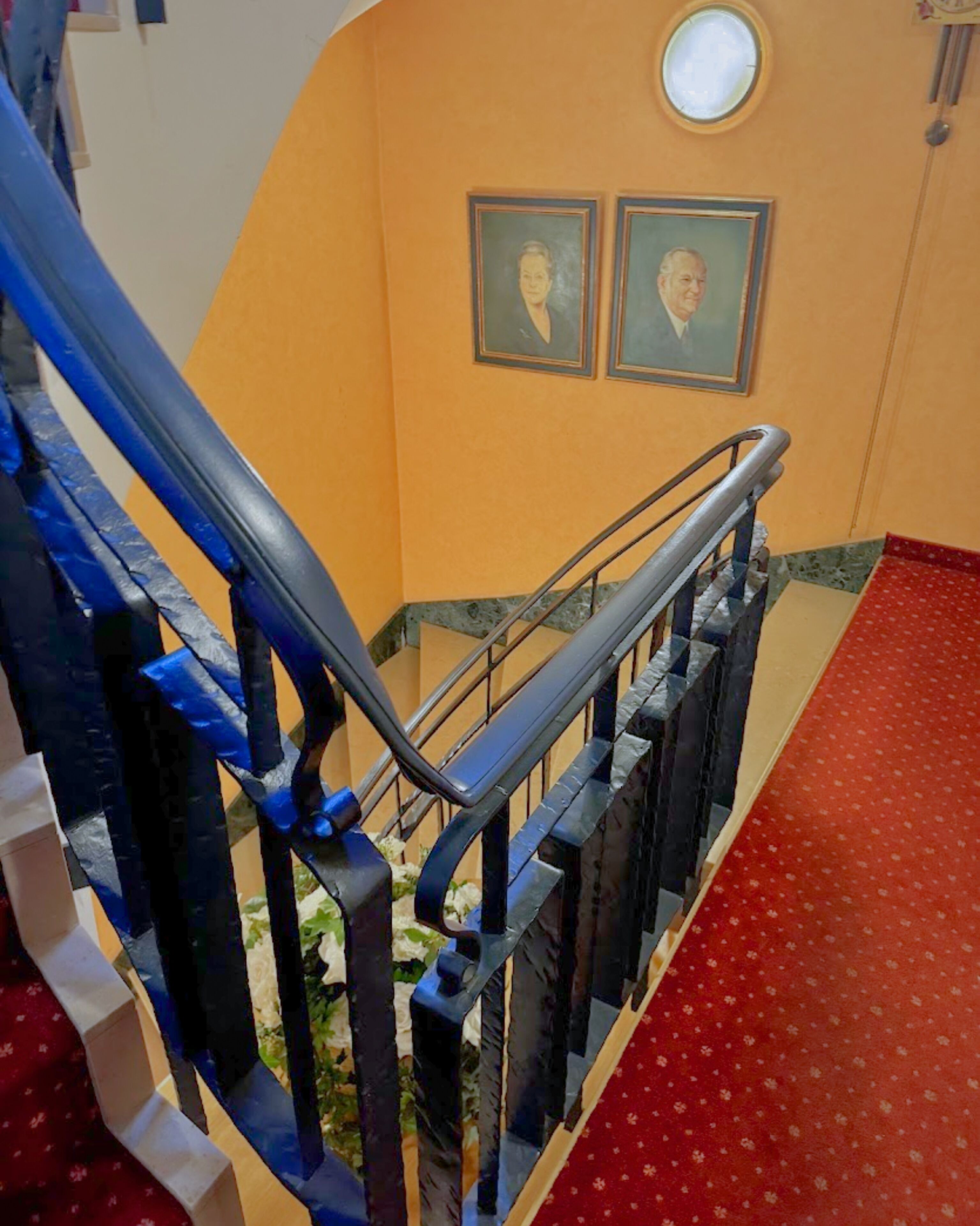 A staircase with metal railings, a red carpeted area, and two framed portraits on the wall.