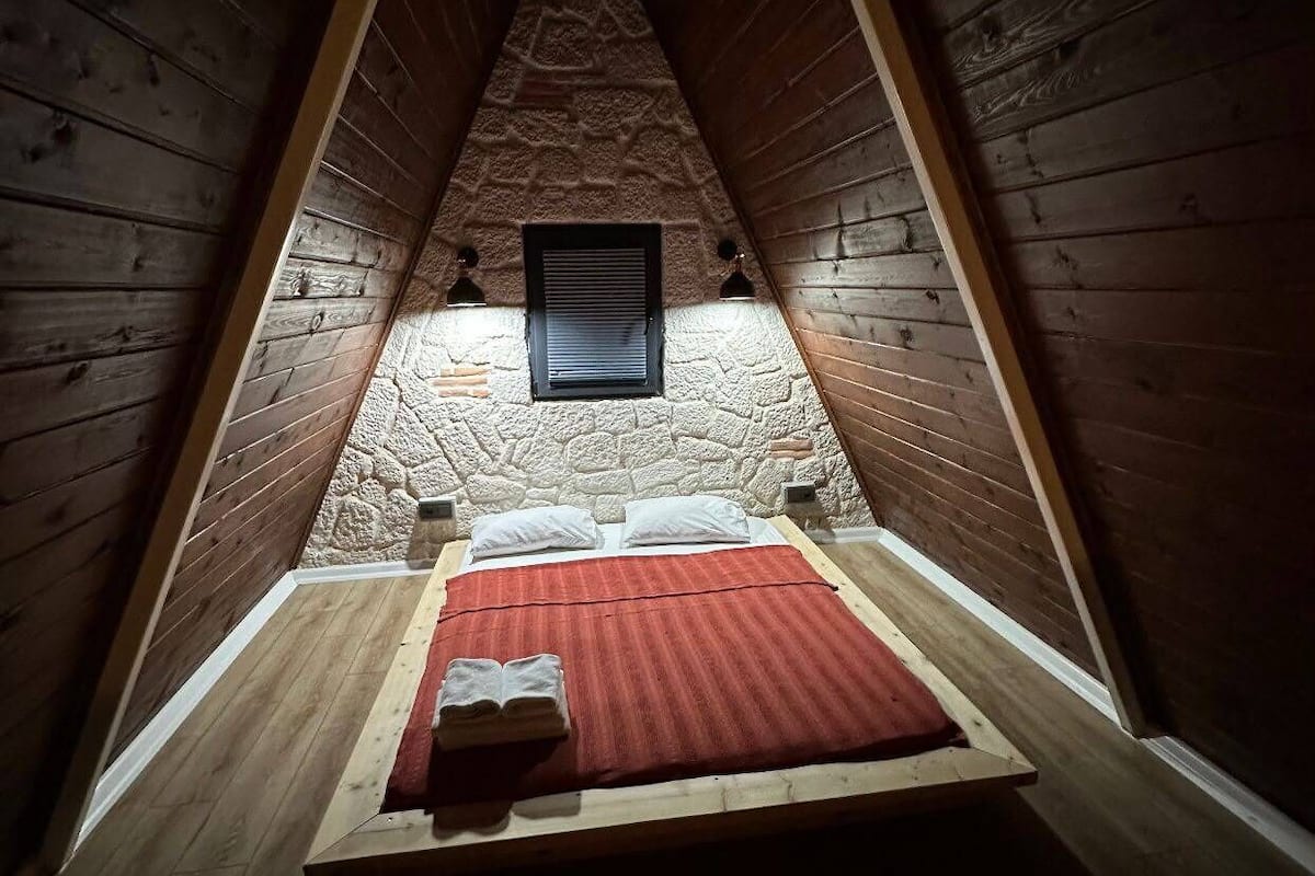 Um quarto de sótão pequeno e acolhedor, com teto inclinado, uma cama de solteiro, uma parede de pedra e duas luminárias.