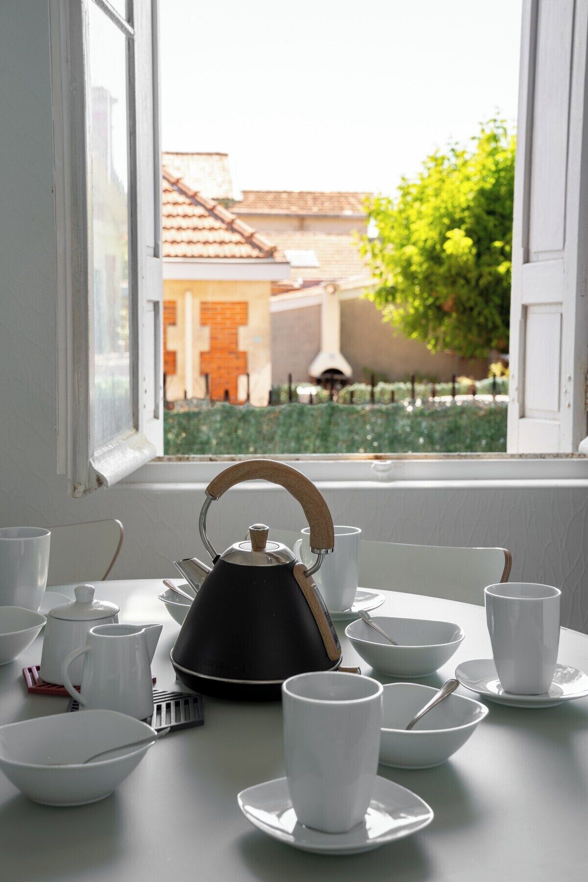 Une table avec une bouilloire, des tasses et des soucoupes, placée près d’une fenêtre donnant sur un bâtiment et de la verdure.