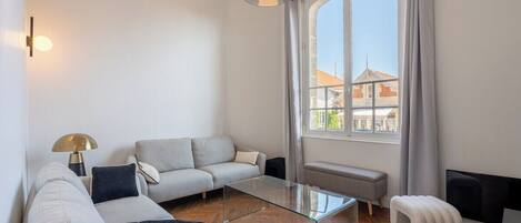 A modern living room with a sofa, glass coffee table, and a view of buildings through a large window.