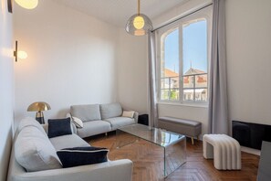 A modern living room with a sofa, glass coffee table, and a view of buildings through a large window. - Villa Le Cid *Charm, Elegance, and the Beaches of Soulac* (Soulac-sur-Mer)