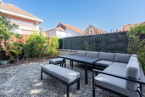 An outdoor patio with a sofa, table, and bench, surrounded by a hedge and a stone wall. - Villa Le Cid *Charm, Elegance, and the Beaches of Soulac* (Soulac-sur-Mer)