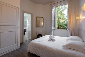 A bedroom with a bed, a window with a view, a mirror, and a door leading to a bathroom. - Villa Le Cid *Charm, Elegance, and the Beaches of Soulac* (Soulac-sur-Mer)