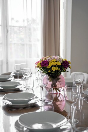 A dining table set with plates and glasses, a pink vase with flowers, and a window with curtains. - Villa Le Cid *Charm, Elegance, and the Beaches of Soulac* (Soulac-sur-Mer)