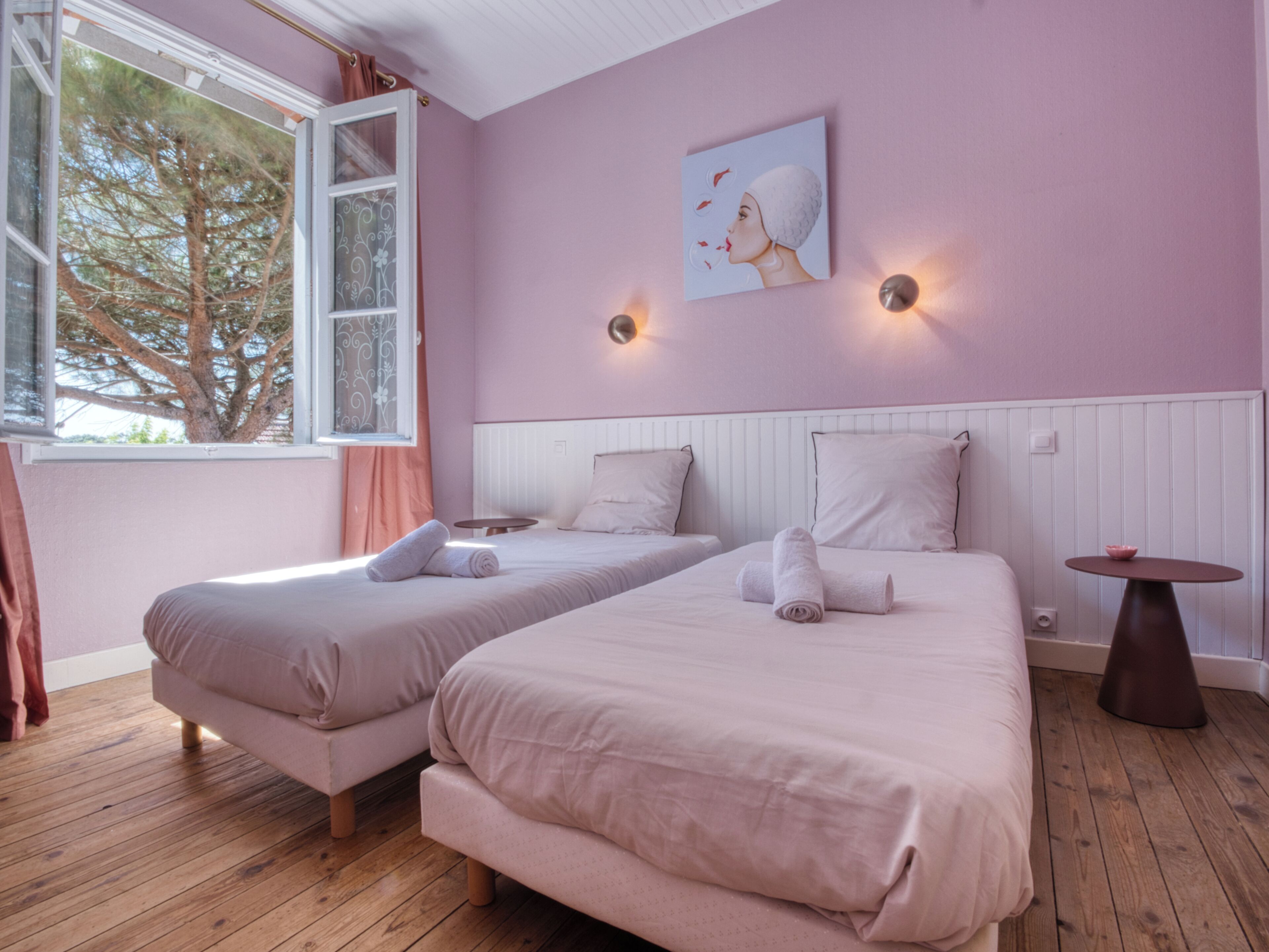 A room with two beds, a window with curtains, a painting on the wall, and a small table.