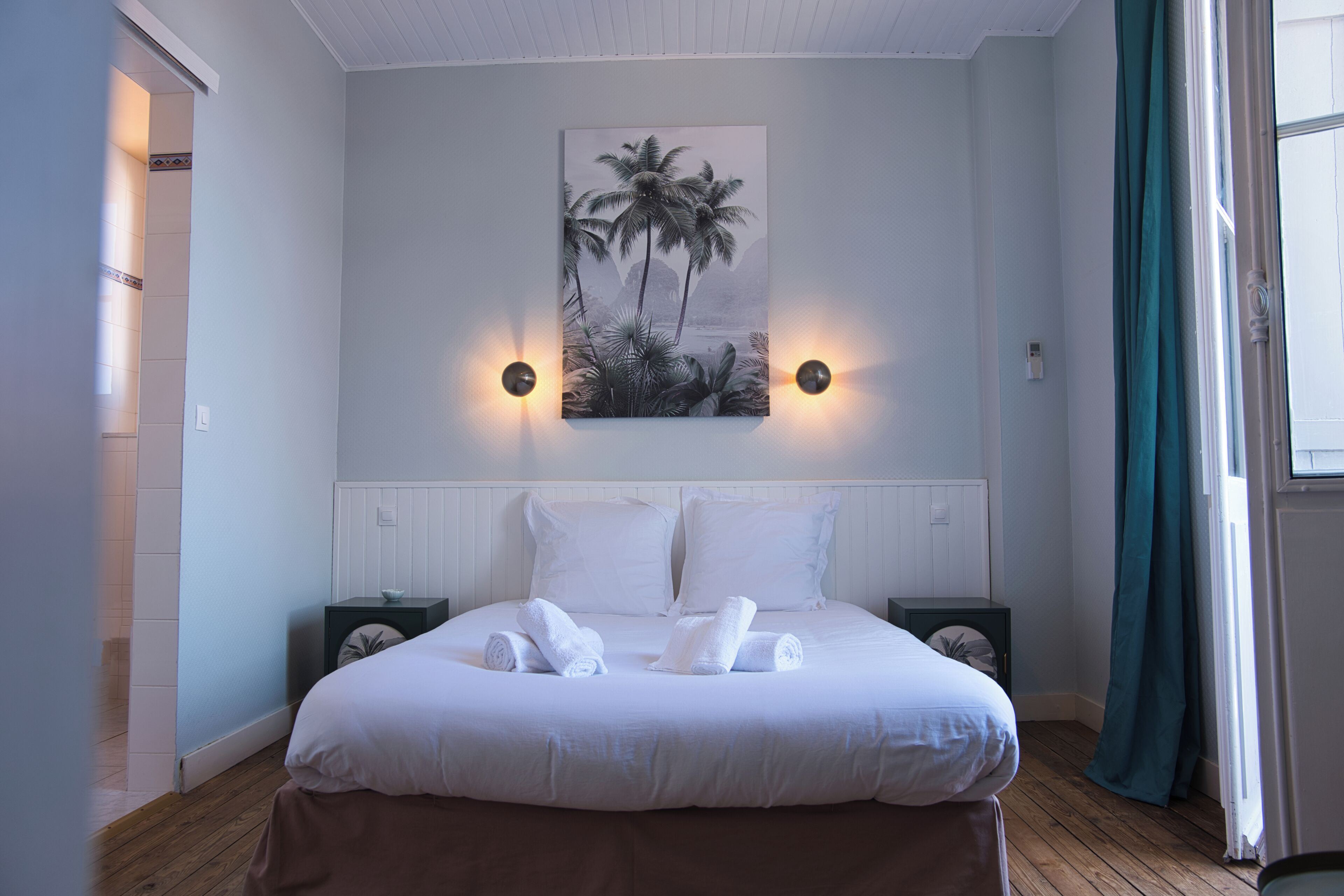 Habitación de hotel con cama, dos mesitas de noche, un cuadro colgado en la pared y una ventana con cortinas.