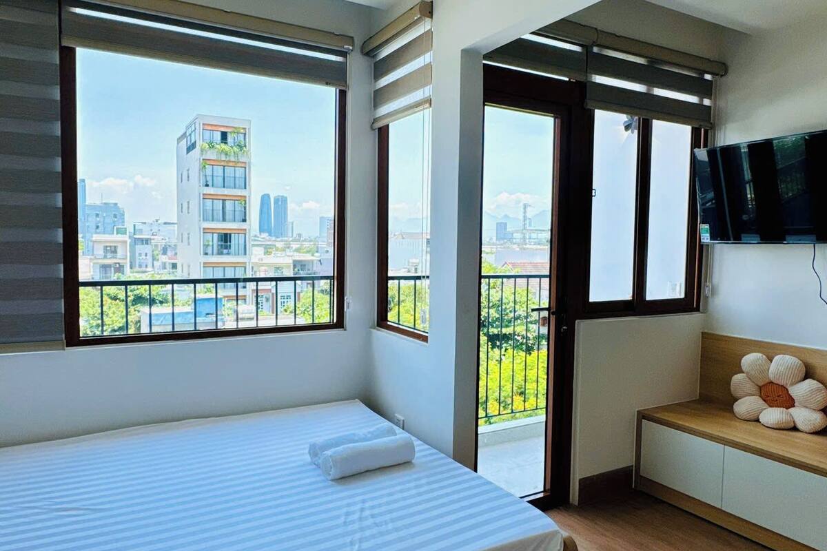 A modern bedroom with a large bed, a TV mounted on the wall, and a balcony with a city view.
