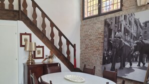 Un coin repas avec une table ronde, des chaises en bois, un escalier et un mur en briques orné d’une grande affiche.