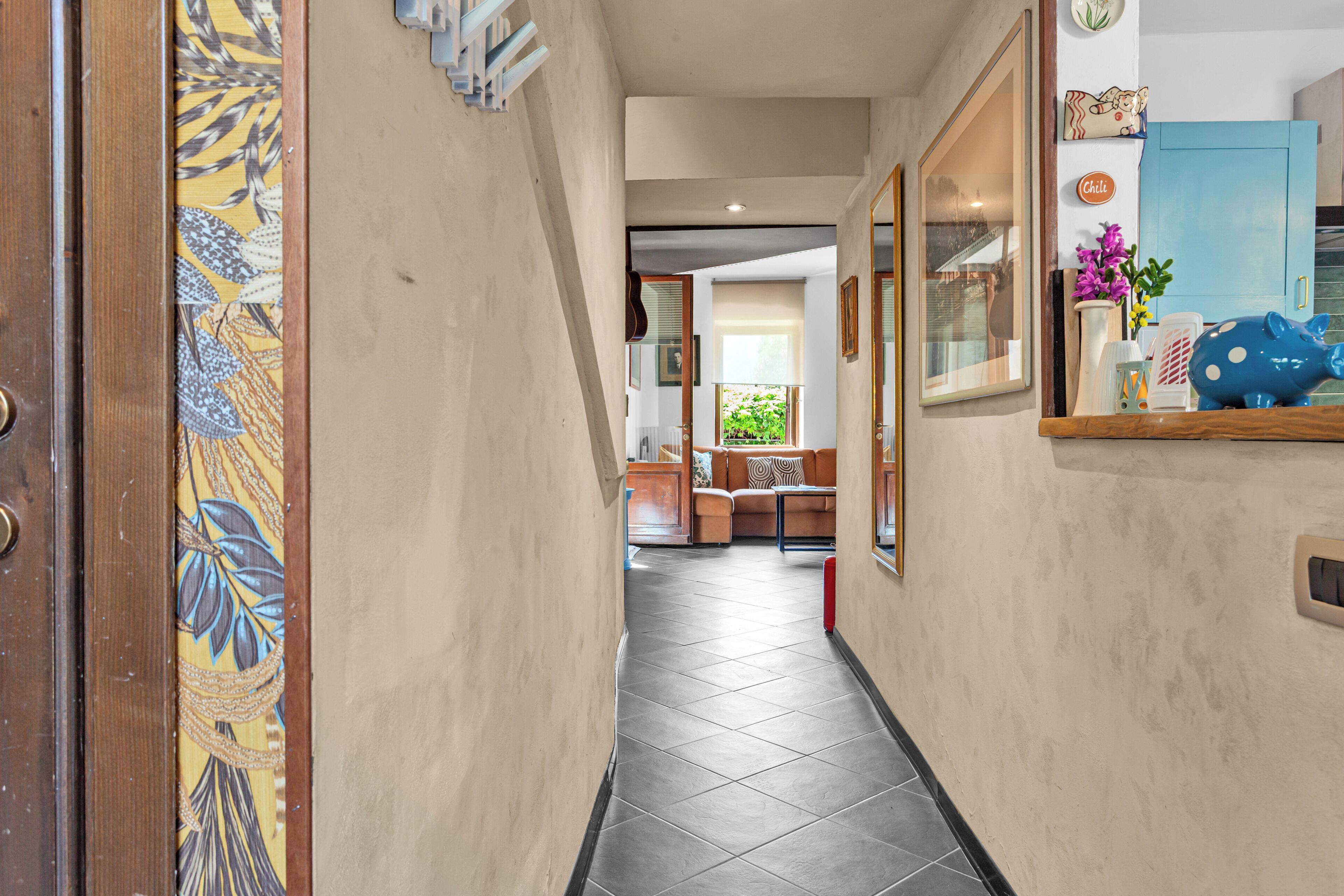 A hallway with a tiled floor, a wooden door with a floral pattern, a blue cabinet, and a window with greenery outside.