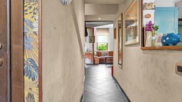 A hallway with a tiled floor, a wooden door with a floral pattern, a blue cabinet, and a window with greenery outside.