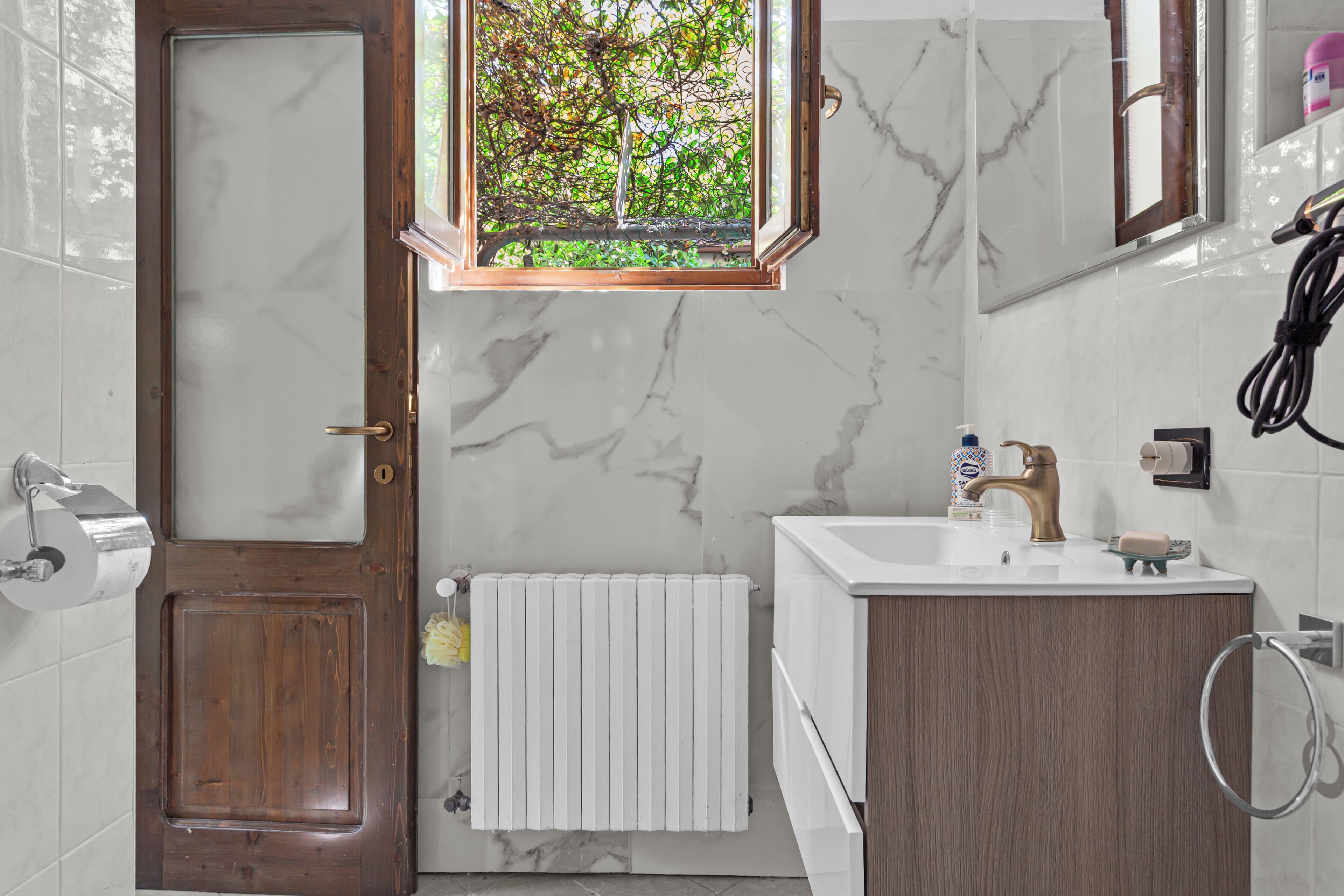 A bathroom with a wooden door, a window with a view of trees, a white radiator, a sink with a mirror, and a towel rack.