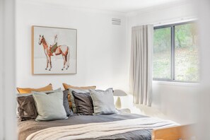 A bedroom with a bed, pillows, a blanket, a window with curtains, and a wall-mounted painting of a horse. - The Hide at Big Hill (Big Hill)