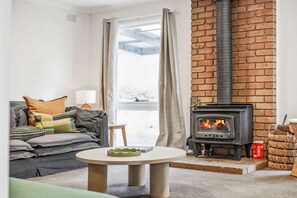 A cozy living room with a fireplace, a sofa, a table, and a view of the outdoors. - The Hide at Big Hill (Big Hill)