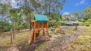 A wooden play structure with a slide and swings, surrounded by trees and a house in the background. - The Hide at Big Hill (Big Hill)
