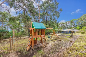 A wooden play structure with a slide and swings, surrounded by trees and a house in the background. - The Hide at Big Hill (Big Hill)