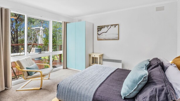 A bedroom with a bed, a chair, a wardrobe, and a window with a view of trees. - The Hide at Big Hill (Big Hill)