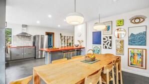 A modern kitchen with stainless steel appliances, a wooden dining table with chairs, and a wall decorated with various artworks. - The Hide at Big Hill (Big Hill)