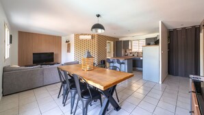Et moderne køkken med en central ø, et spiseområde med et træbord og stole, samt en opholdsstue med sofa og fjernsyn.