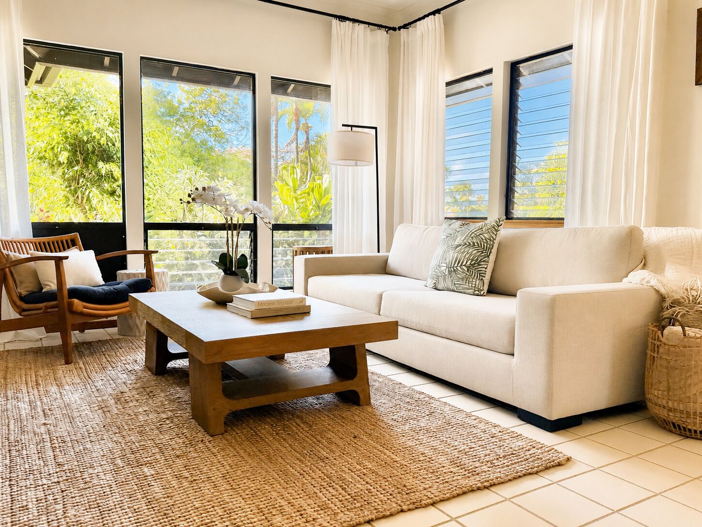 A living room with a sofa, coffee table, and chair, large windows with curtains, and a view of greenery outside.