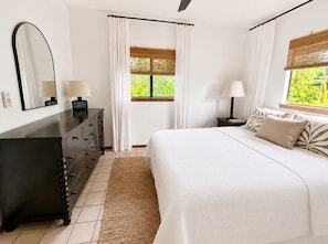 Ein Schlafzimmer mit Bett, Kommode, einem Fenster mit Vorhängen und einem Spiegel.