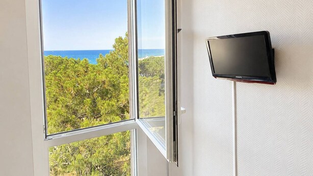 A room with a large window offering a view of the ocean and greenery, a flat-screen TV mounted on the wall. - Charming T2 with sea view - 2 guests (Argelès-sur-Mer)