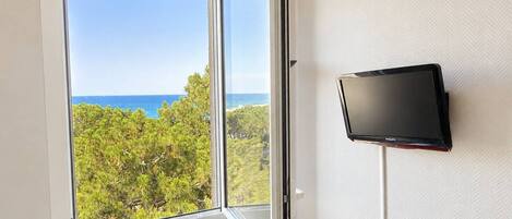 A room with a large window offering a view of the ocean and greenery, a flat-screen TV mounted on the wall.