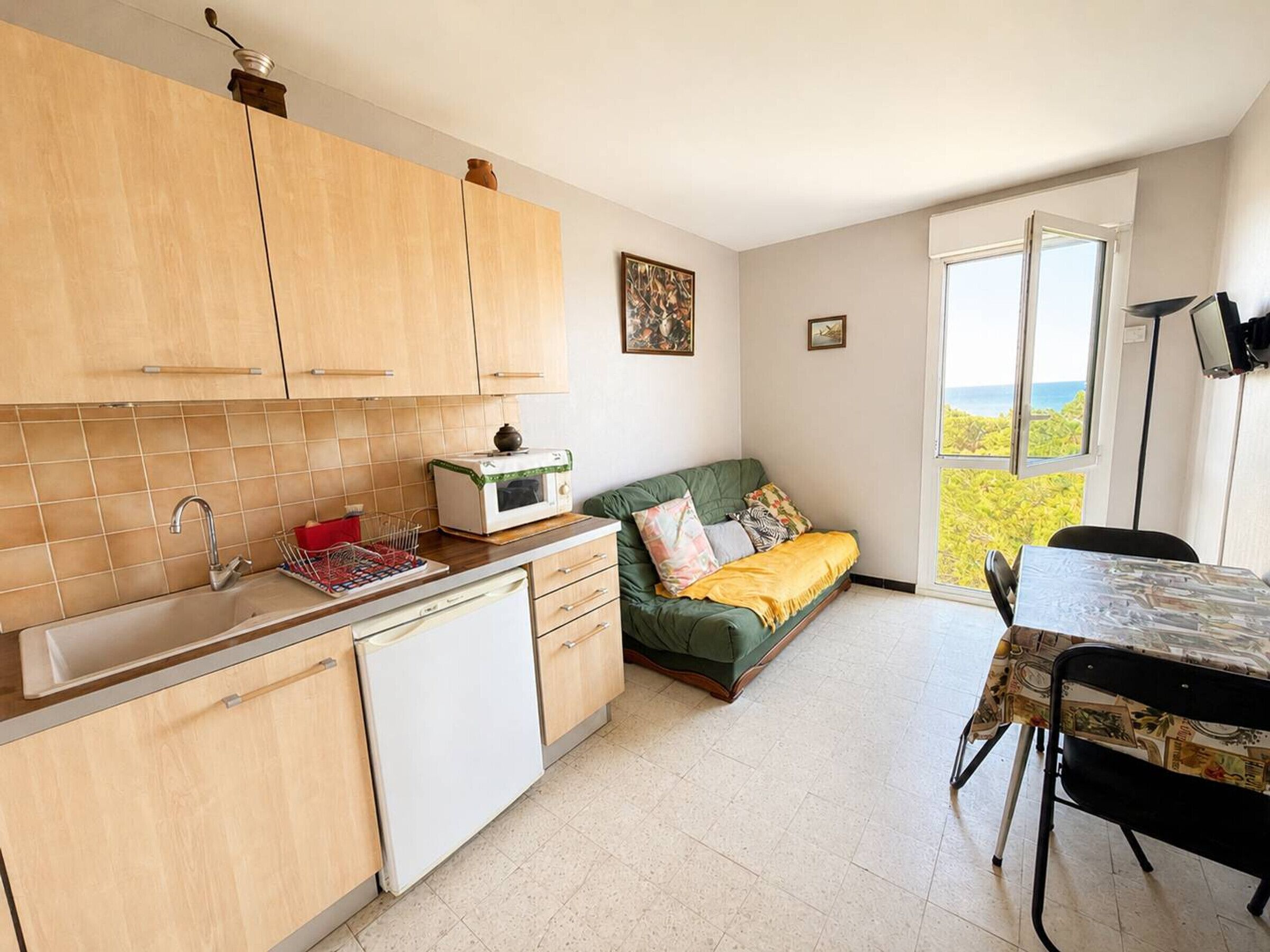 Cocina compacta con lavamanos, microondas y armarios de madera. Hay un sofá verde con una manta y cojines amarillos. Una mesa de comedor con sillas se encuentra cerca de un ventanal con vistas.