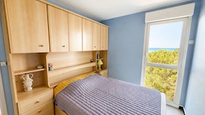 A bedroom with a bed, wooden cabinets, a nightstand, a window with a view of greenery, and a door. - Charming T2 with sea view - 2 guests (Argelès-sur-Mer)