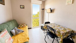A room with a green couch, a yellow blanket, a table with a tablecloth, a chair, a lamp, and a view through a window. - Charming T2 with sea view - 2 guests (Argelès-sur-Mer)
