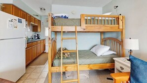 Una cama litera con escalera, un escritorio, una lámpara y una zona de cocina con armarios y un refrigerador.