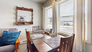 Un comedor con una mesa de madera, sillas, un cojín azul, un estante de madera con objetos y una ventana con cortinas.