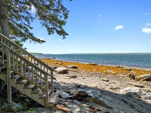 A wooden staircase leading to a rocky shoreline with a view of the water and distant land. - Charming Home With Stunning bay Views, Sunroom, Gazebo, Deck, & Grill (Trenton)