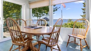 Um alpendre coberto com mesa e cadeiras de madeira, janelas amplas e vista para a água.