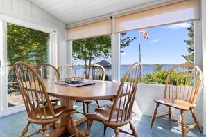 A covered patio with a wooden table and chairs, large windows, and a view of the water. - Charming Home With Stunning bay Views, Sunroom, Gazebo, Deck, & Grill (Trenton)