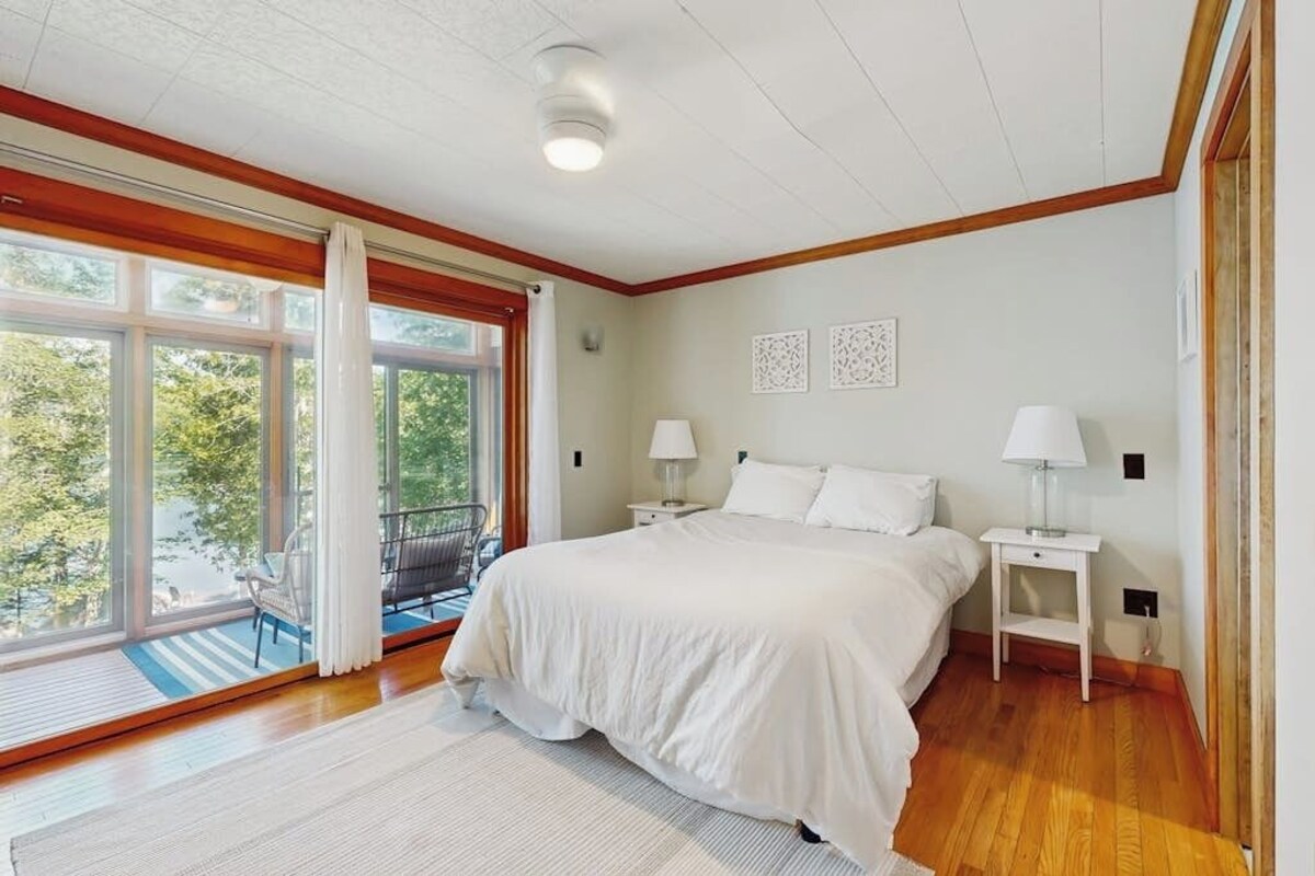 A bedroom with a large bed, two bedside tables with lamps, a window with curtains, and a view of greenery outside.