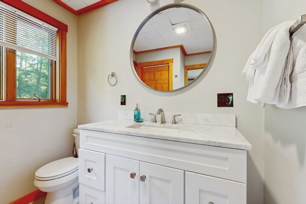 A bathroom with a white vanity, a round mirror, a toilet, and a window with blinds.