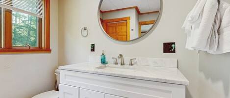 A bathroom with a white vanity, a round mirror, a toilet, and a window with blinds.