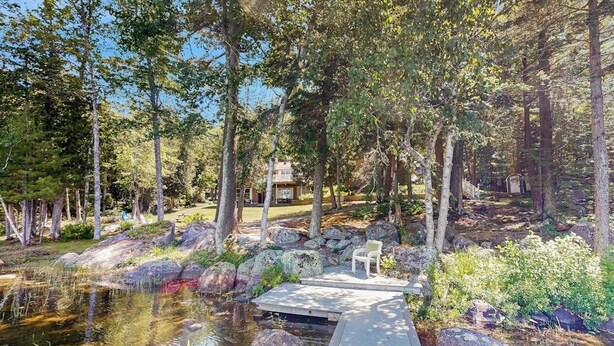 Un cadre extérieur paisible avec un étang, une terrasse en bois et une maison entourée d’arbres.